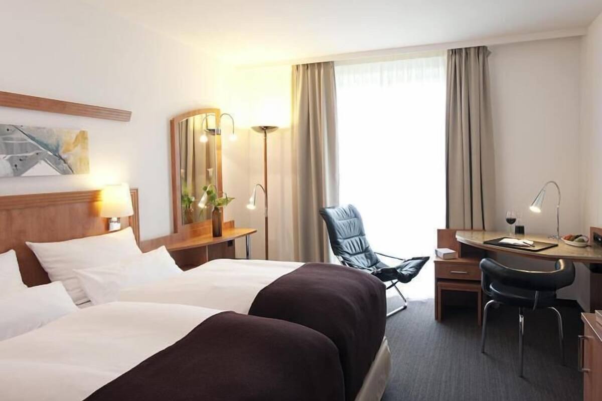A hotel room with two beds, a desk, a chair, a lamp, a mirror, and a window with curtains.