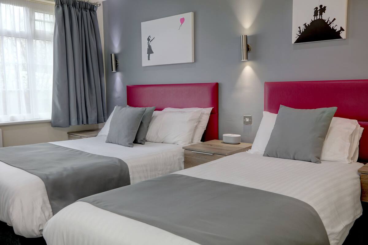 Ein Hotelzimmer mit zwei Betten, grau-weißer Bettwäsche, einem roten Kopfteil, einem Nachttisch mit Lampe und Kunstwerken an den Wänden.