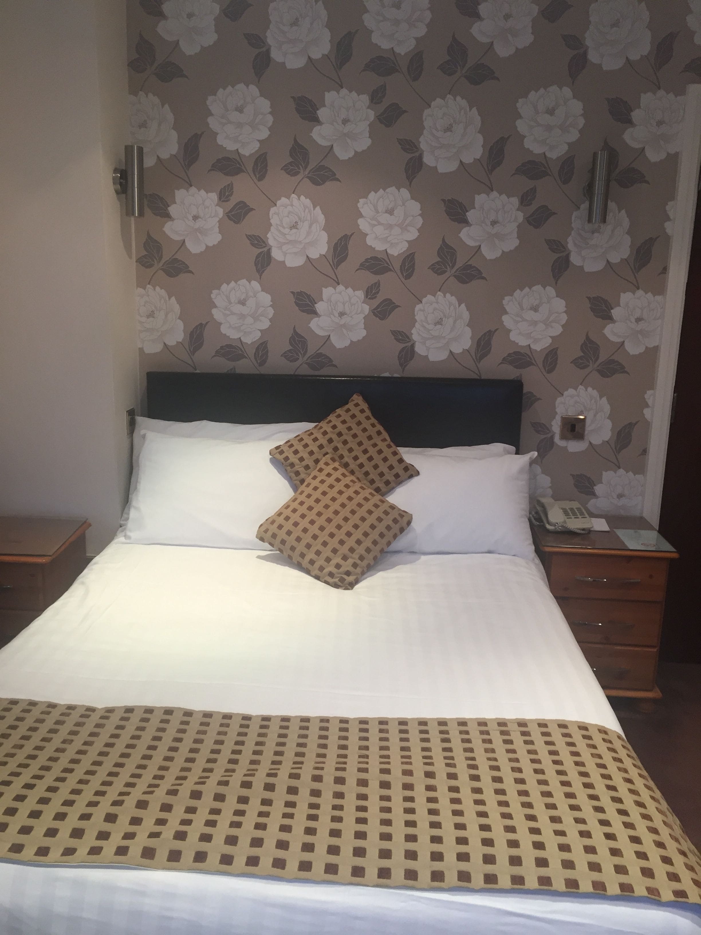 Een hotelkamer met een groot bed, een beddengoed met een patroon, een hoofdeinde, twee nachtkastjes en bloemenbehang.