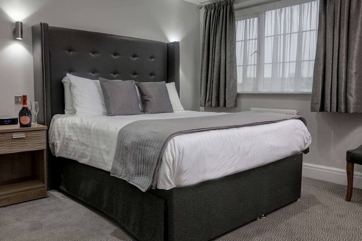 Ein modernes Schlafzimmer mit einem gepolsterten Kopfteil, einem großen Bett mit weißer und grauer Bettwäsche, einem Nachttisch mit Flasche und Gläsern sowie einem Fenster mit Vorhängen.