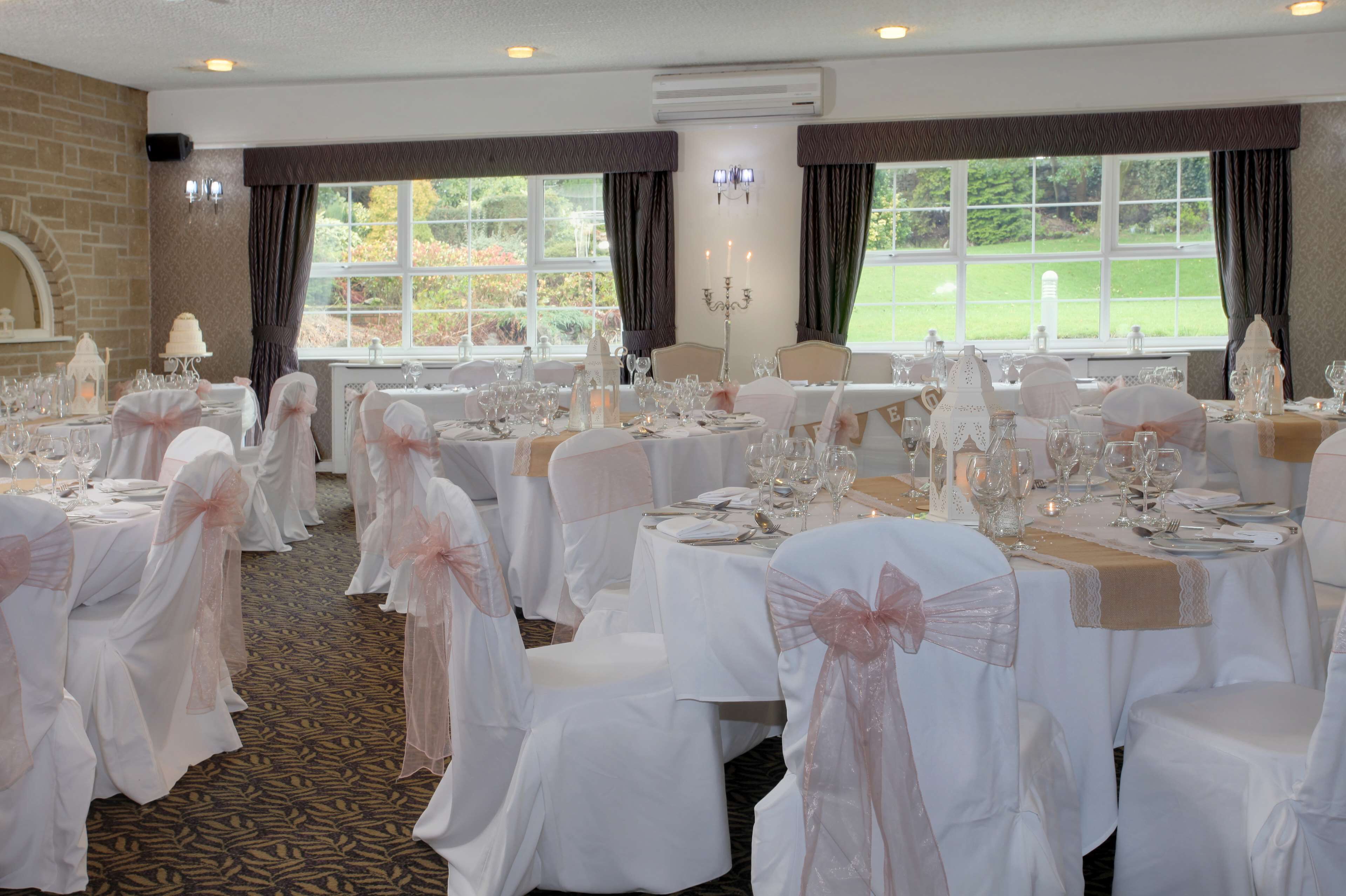 Un salón de banquetes con mesas redondas dispuestas para un evento formal, manteles blancos y sillas adornadas con cintas rosas.