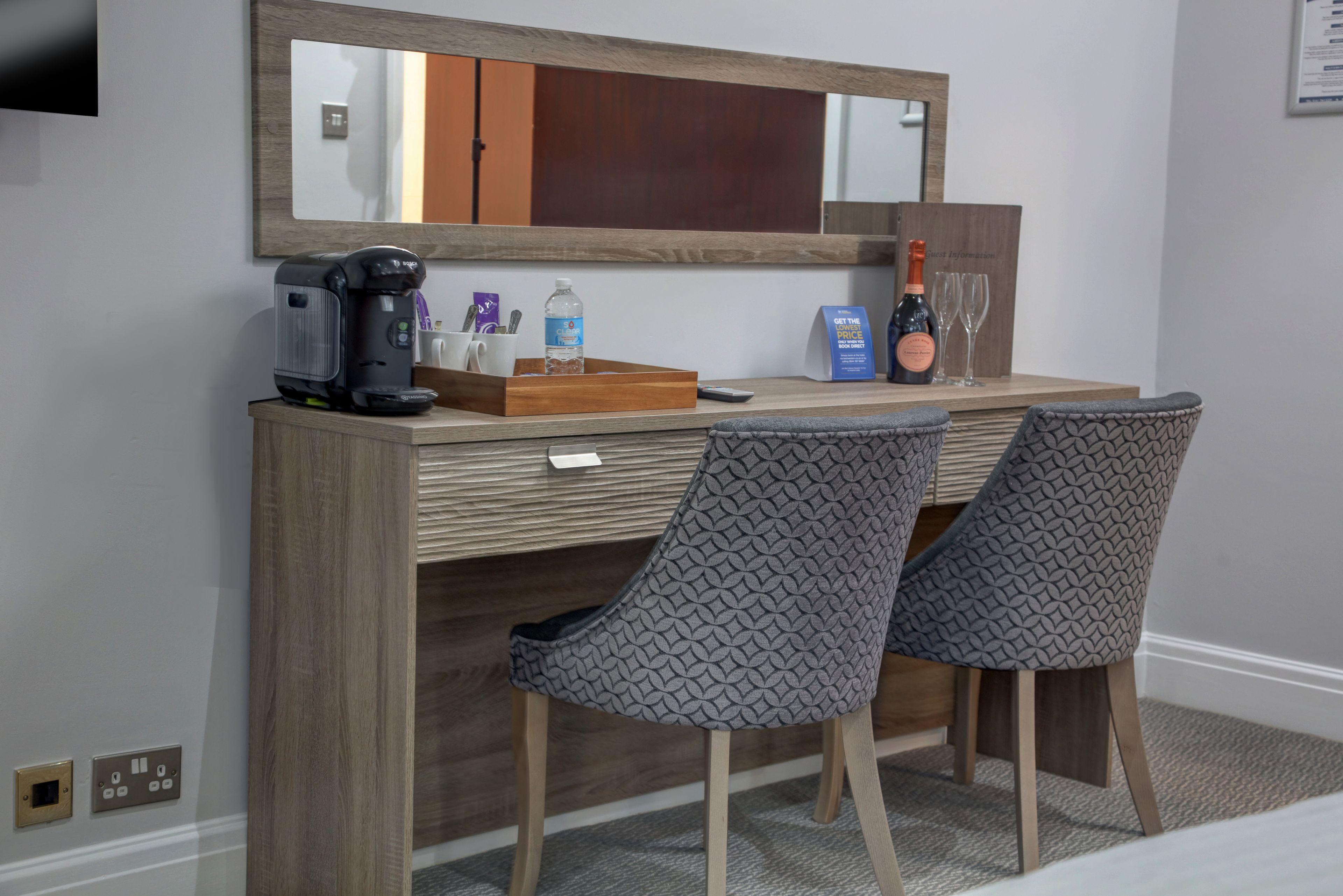 A hotel room with a wooden desk, two patterned chairs, a coffee maker, a bottle of water, a bottle of wine, and a mirror.