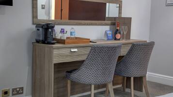 A hotel room with a wooden desk, two patterned chairs, a coffee maker, a bottle of water, a bottle of wine, and a mirror.