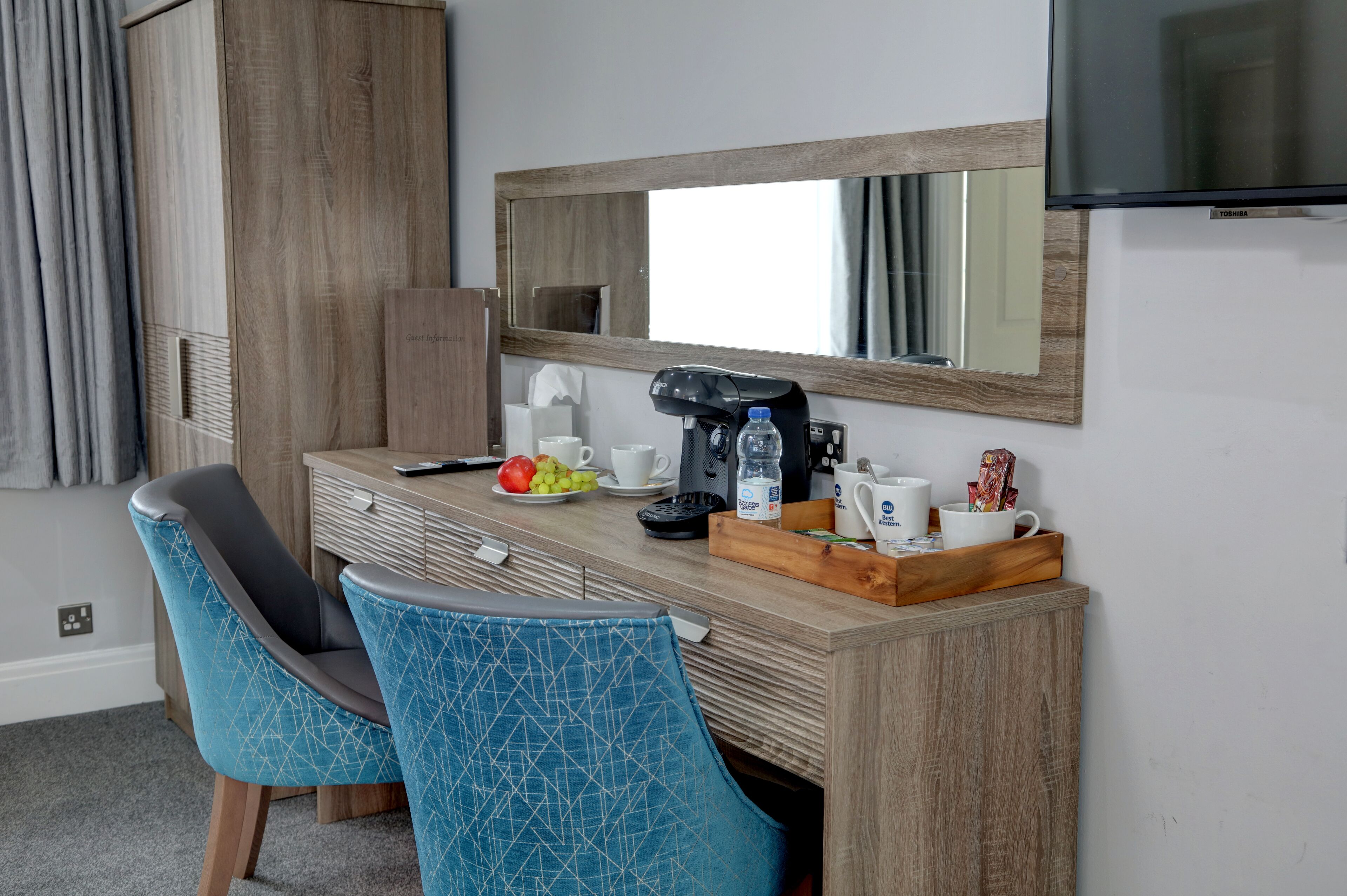Habitación de hotel con un escritorio de madera, dos sillas azules con estampado, una cafetera, una botella de agua, una taza, una bandeja con una mug y un televisor en la pared.