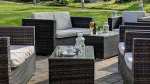 Mobiliario de mimbre exterior con cojines, una mesa de vidrio con tazas y una copa de vino, y un jardín al fondo.