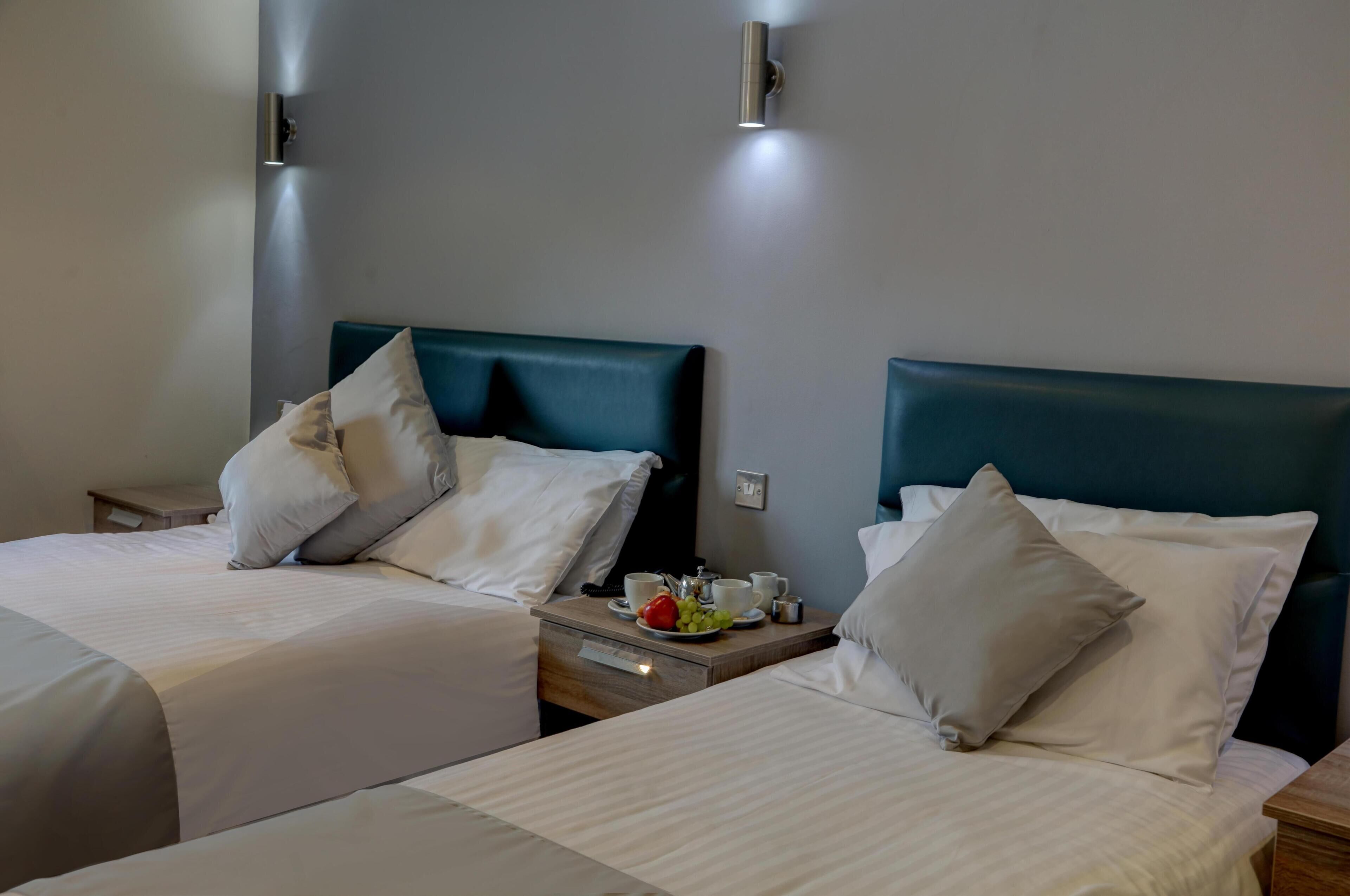 A hotel room with two beds, each with a headboard, a bedside table with a lamp, and a small table with a fruit bowl and cups.