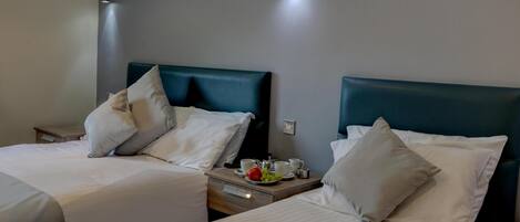 A hotel room with two beds, each with a headboard, a bedside table with a lamp, and a small table with a fruit bowl and cups.