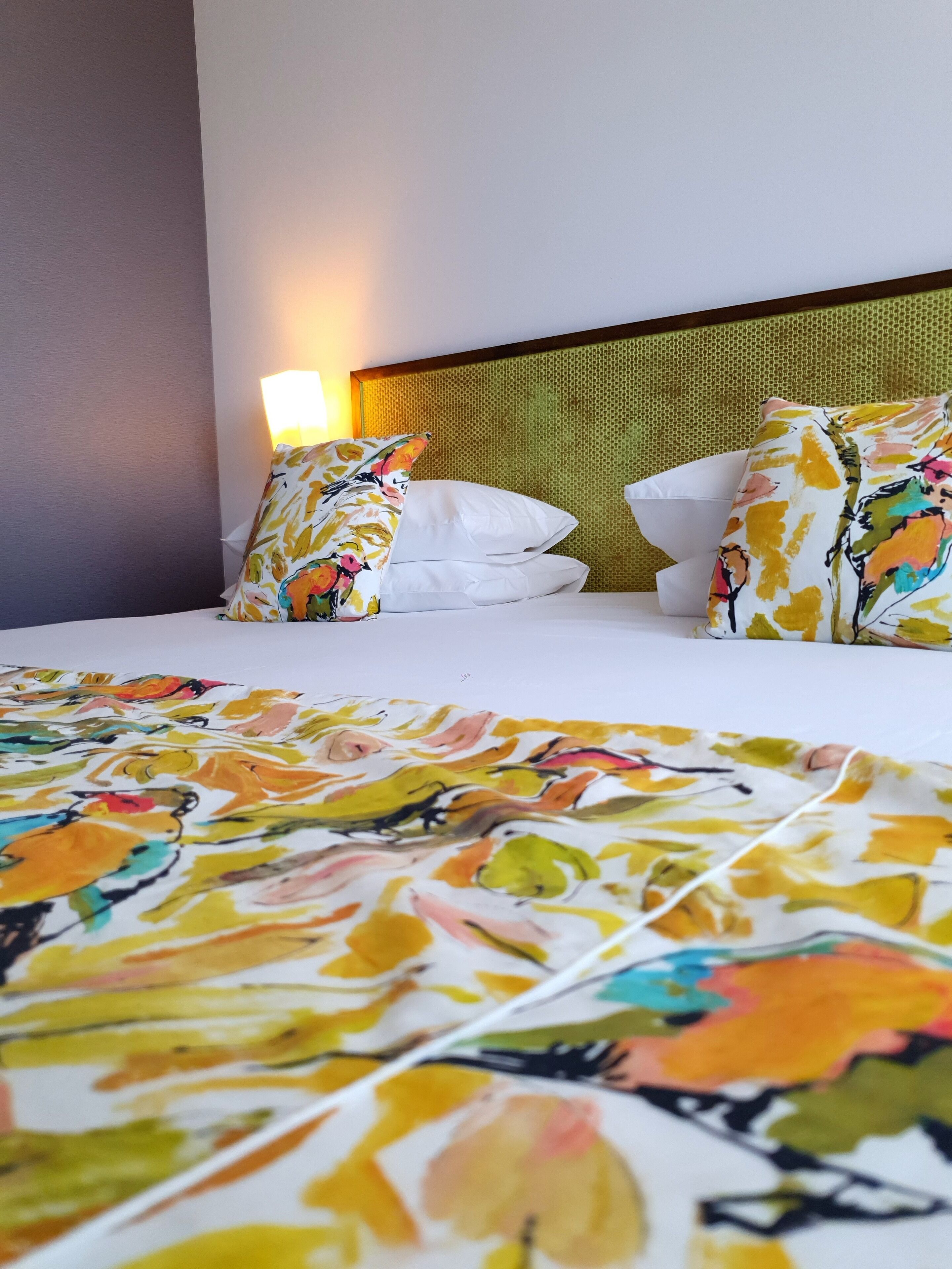 Une chambre d’hôtel avec un lit, des oreillers blancs, une couvre-lit à motifs floraux colorés et une tête de lit à texture particulière.