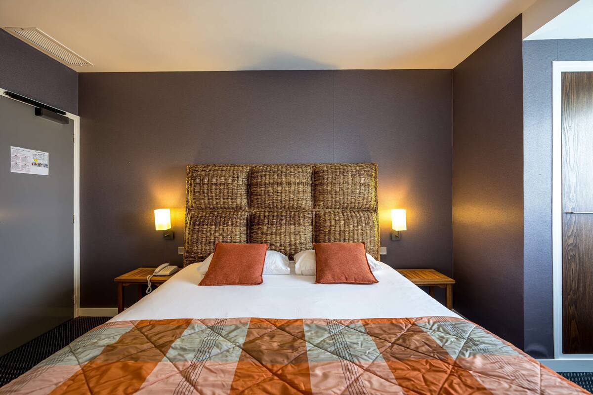 A hotel room with a large bed, a headboard with a woven design, two bedside tables with lamps, and a quilted bedspread.