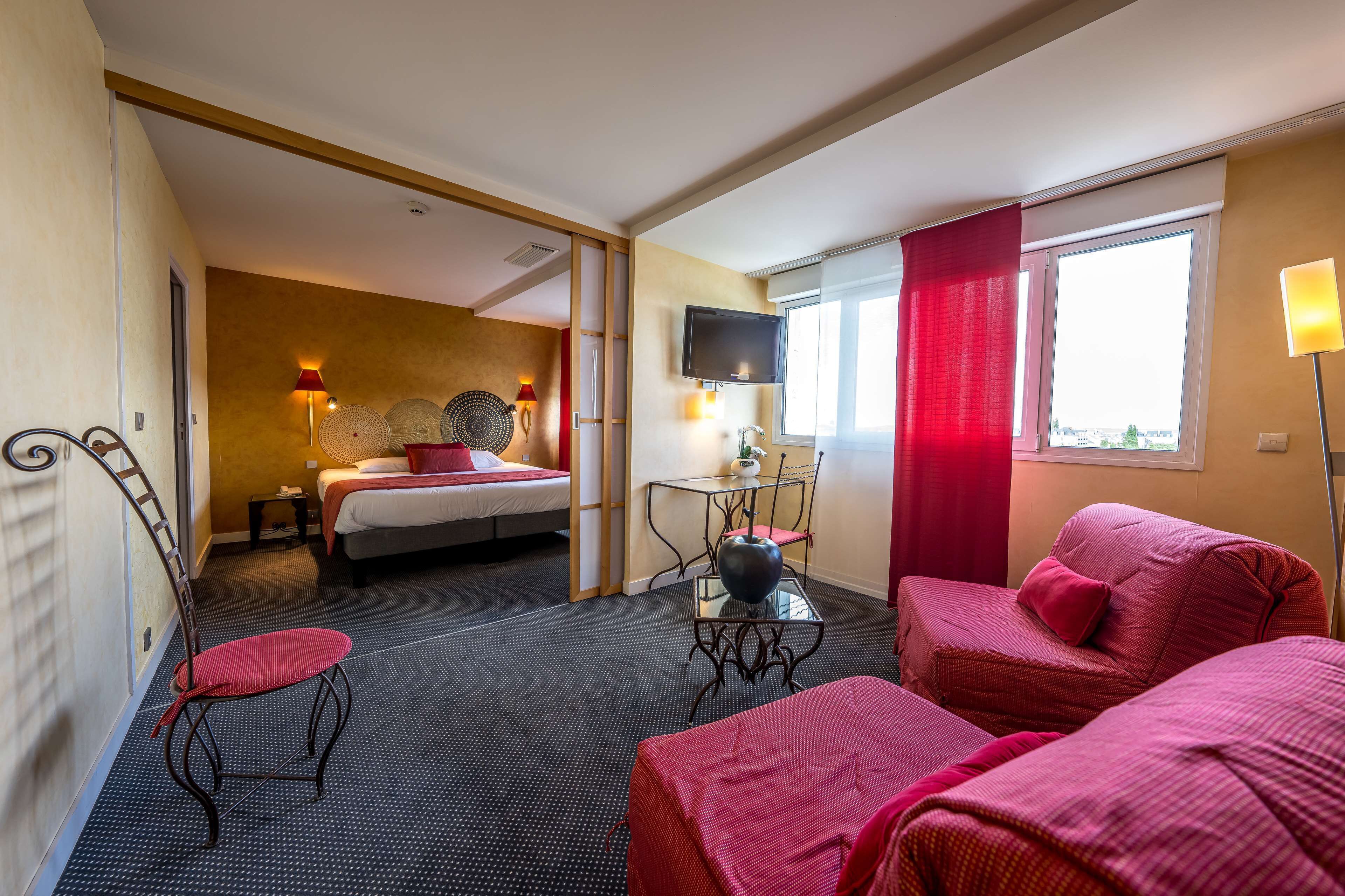 A hotel room with a bed, a sofa, a dining table with chairs, a TV mounted on the wall, and a window with curtains.