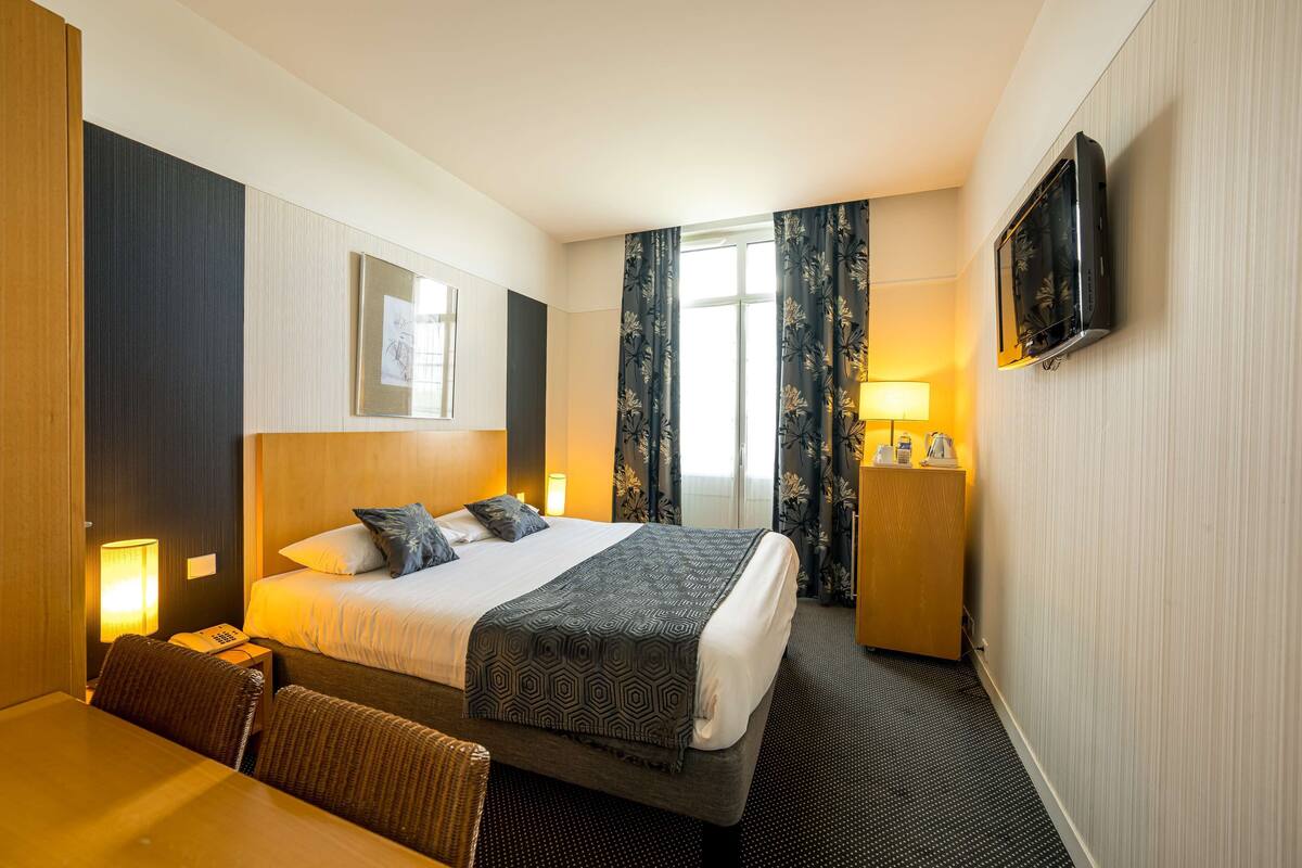 A hotel room with a bed, bedside table, lamp, TV, and a desk with a chair.