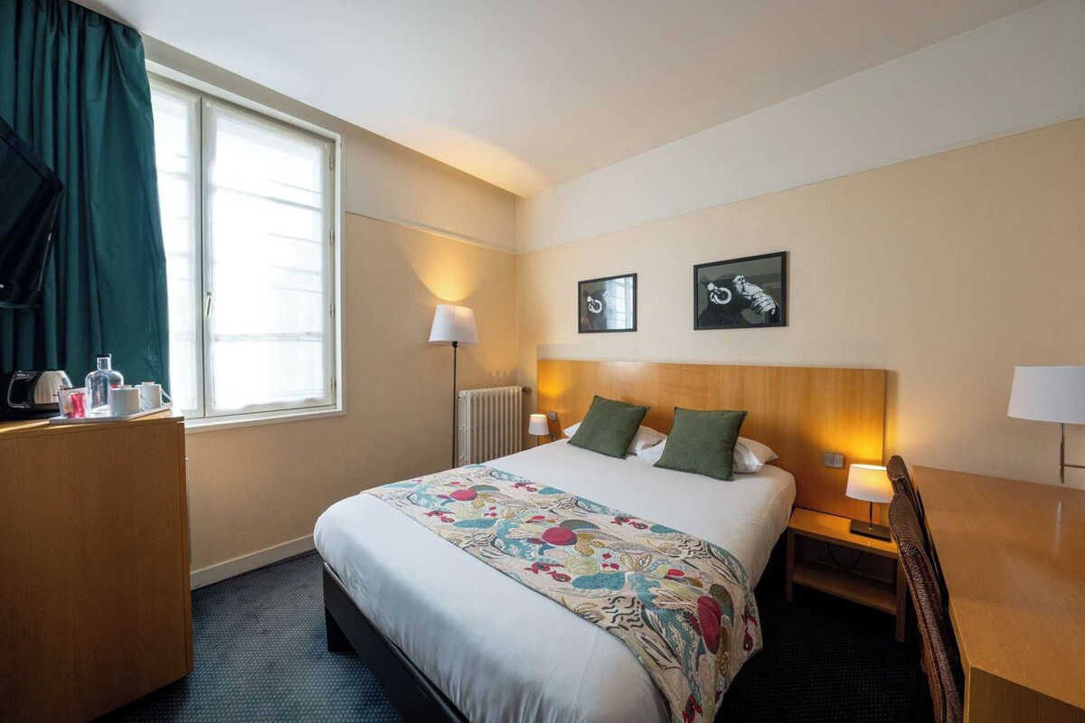 A hotel room with a large bed, bedside lamps, a desk with a chair, a window with curtains, and two wall-mounted artworks.