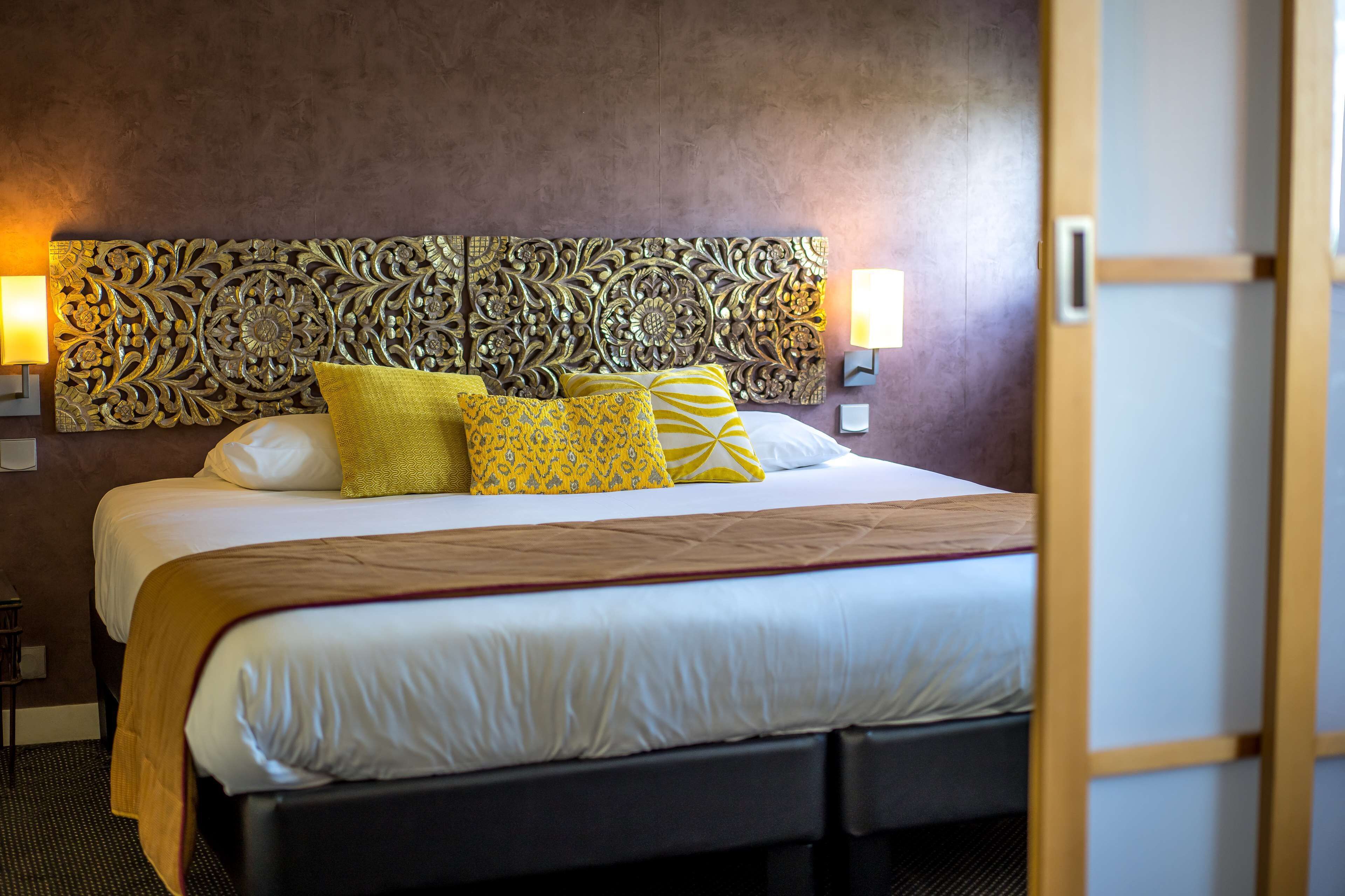 A hotel room with a large bed, decorative headboard, two bedside lamps, and a wooden door.