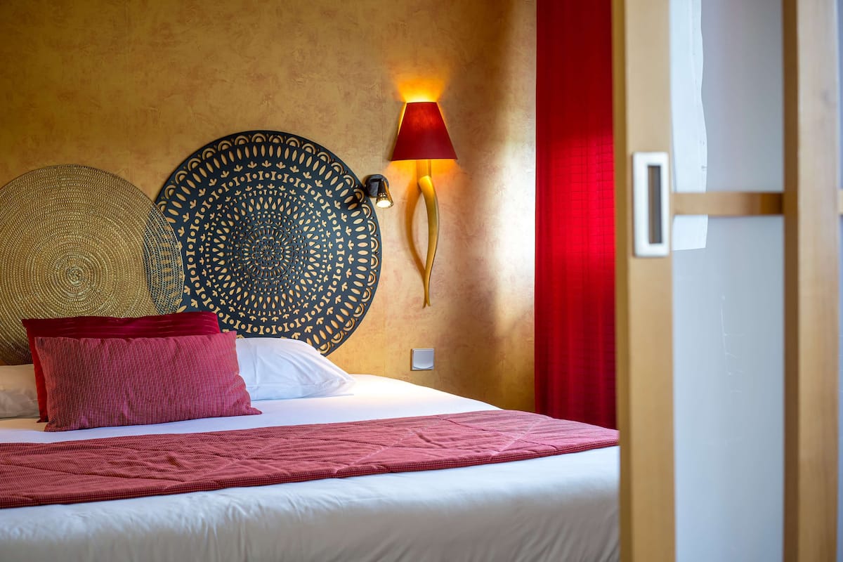 A neatly made bed with a red blanket and pillows, a wall-mounted lamp, and decorative wall hangings.