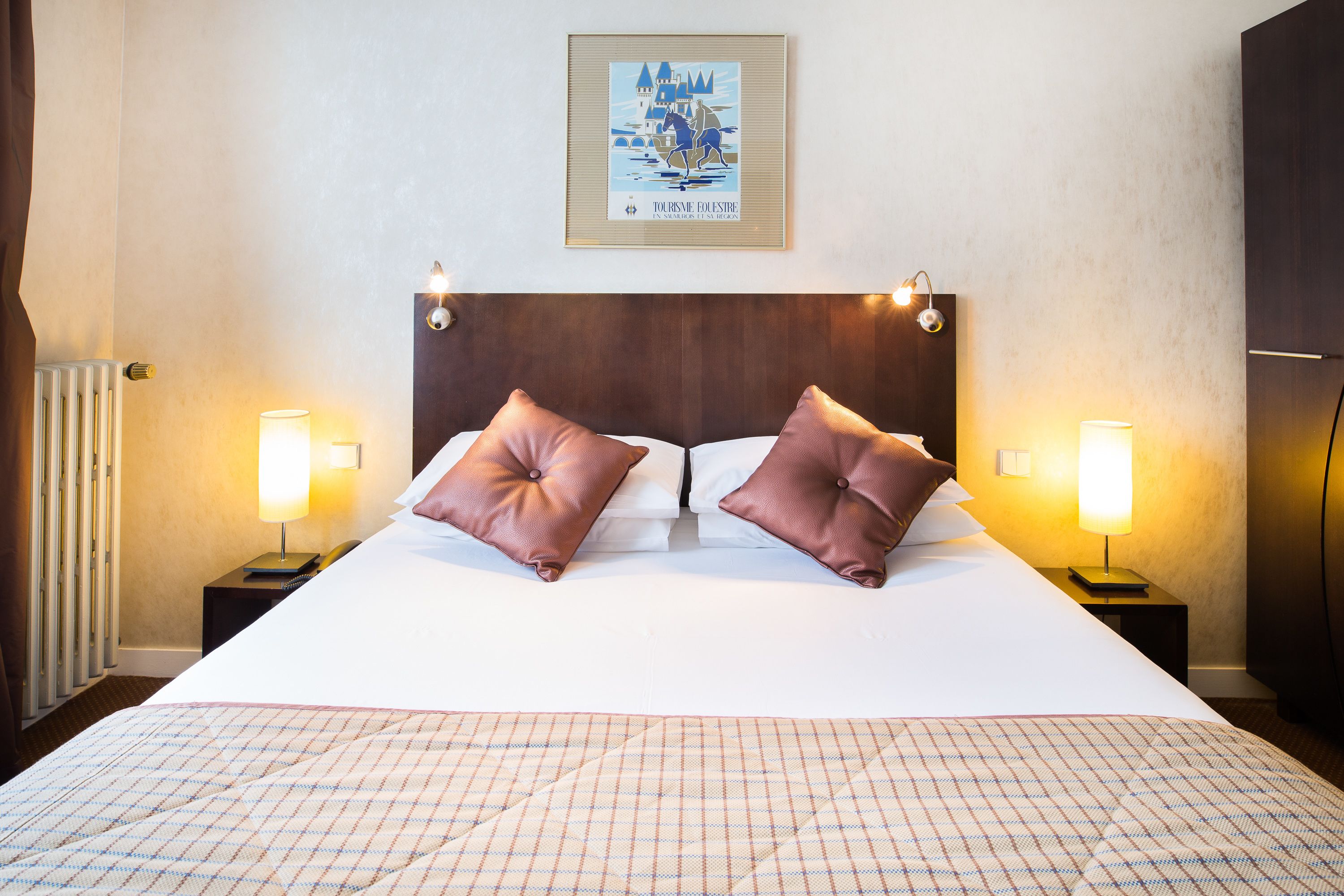 A hotel room with a large bed, two bedside lamps, a framed picture on the wall, and a radiator on the left.