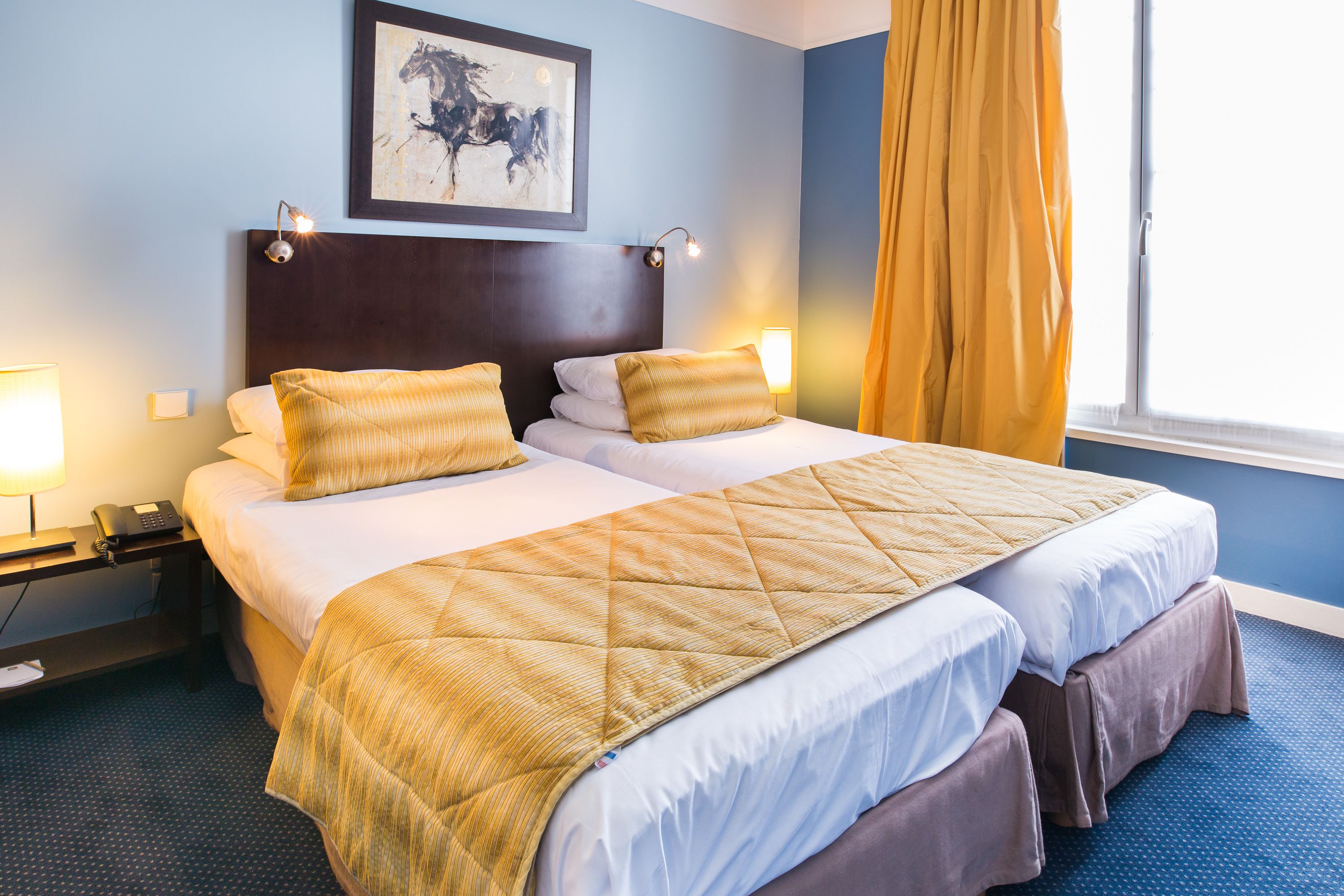 A hotel room with two beds, a headboard, bedside lamps, a painting, and a window with curtains.