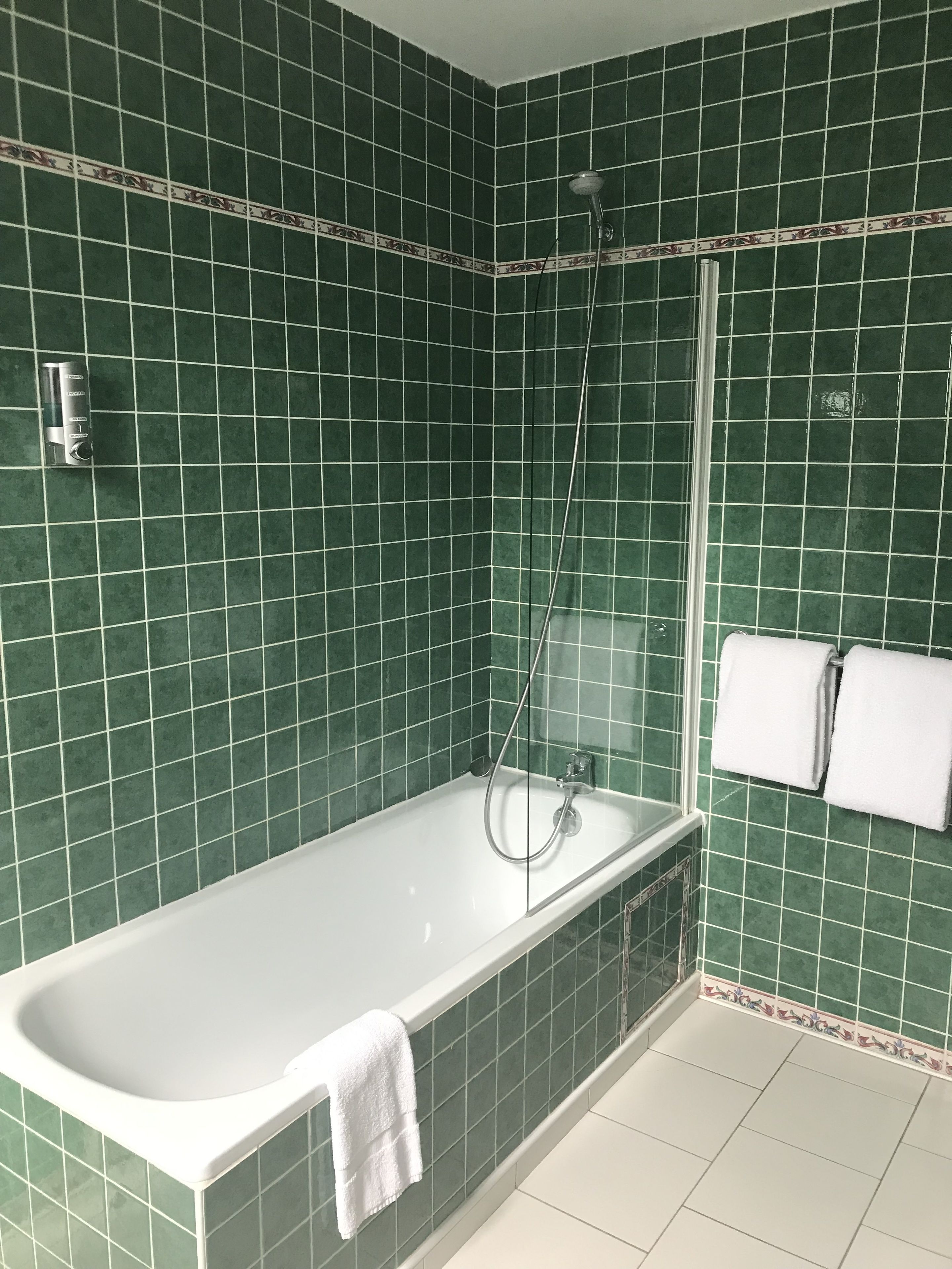 A bathroom with green tiled walls, a white bathtub, and a shower. There are two white towels hanging on the wall.