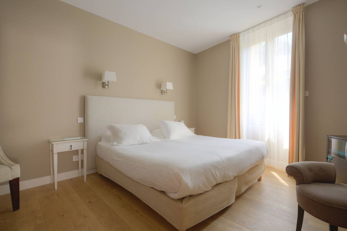 Een slaapkamer met een groot bed, een nachtkastje, een stoel en een raam met gordijnen.