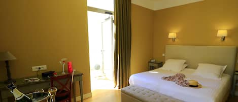 Una habitación de hotel con cama, un escritorio con una botella de champán, una silla y una mesa con un vaso y un objeto rojo.
