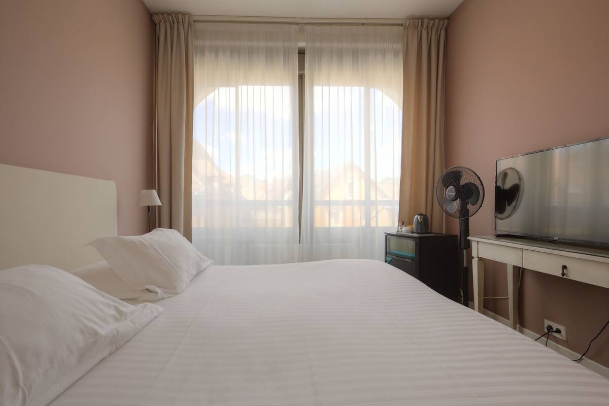 Een hotelkamer met een groot bed, een televisie, een bureau met een ventilator en een raam met dunne gordijnen.