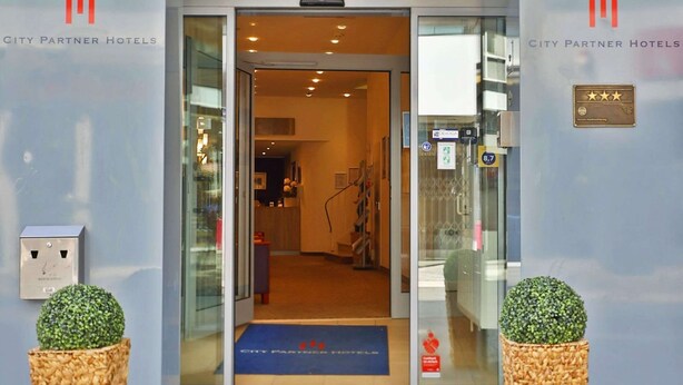 Ingången till Central Hotel i Garmi har en modern glasfasad, en reception som syns genom dörröppningen och två dekorativa blomkrukor på var sin sida.