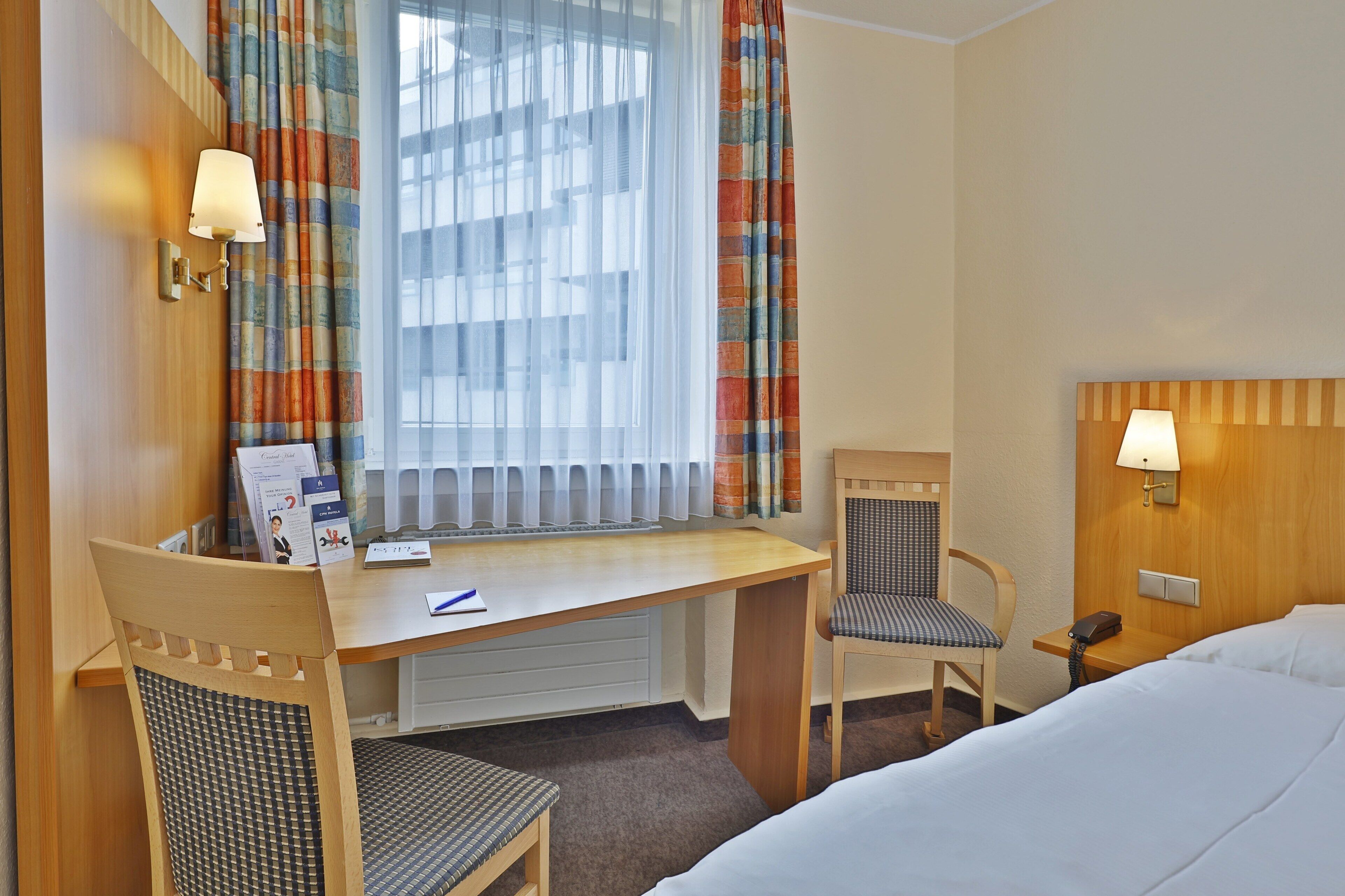 A hotel room with a bed, a desk with a chair, a telephone, and a window with curtains.