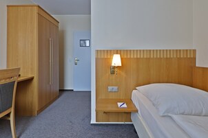 Habitación de hotel con armario de madera, escritorio y silla, mesita de noche con lámpara y cama con ropa blanca.