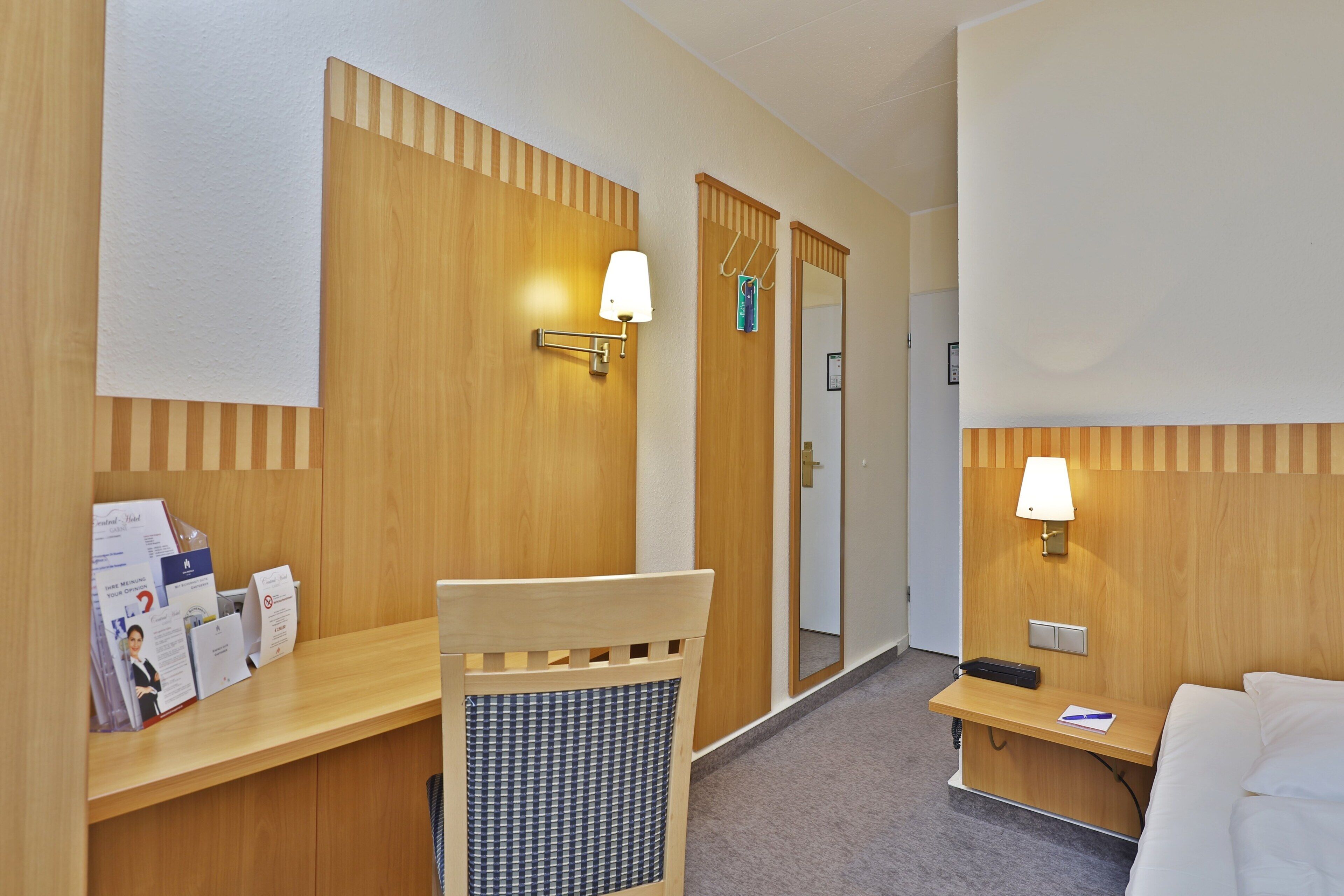 Habitación de hotel con mostrador de recepción de madera, una silla, una mesita de noche con lámpara y una cama.
