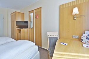 A hotel room with a bed, a desk with a lamp, a chair, and a TV.