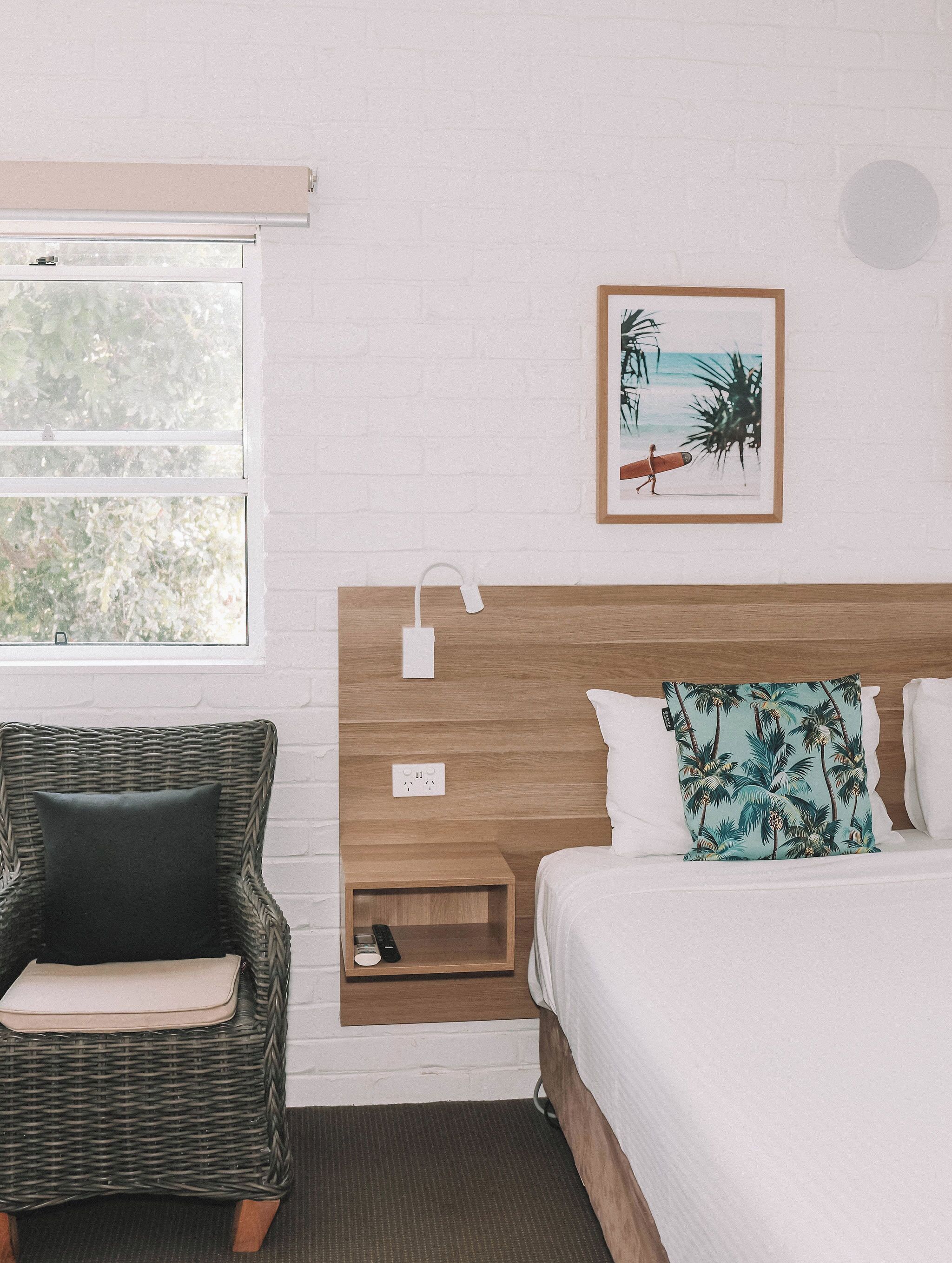 Una habitación de hotel con una cama, una mesita de noche, una silla de mimbre, una lámpara de pared, un enchufe y un cuadro en la pared.