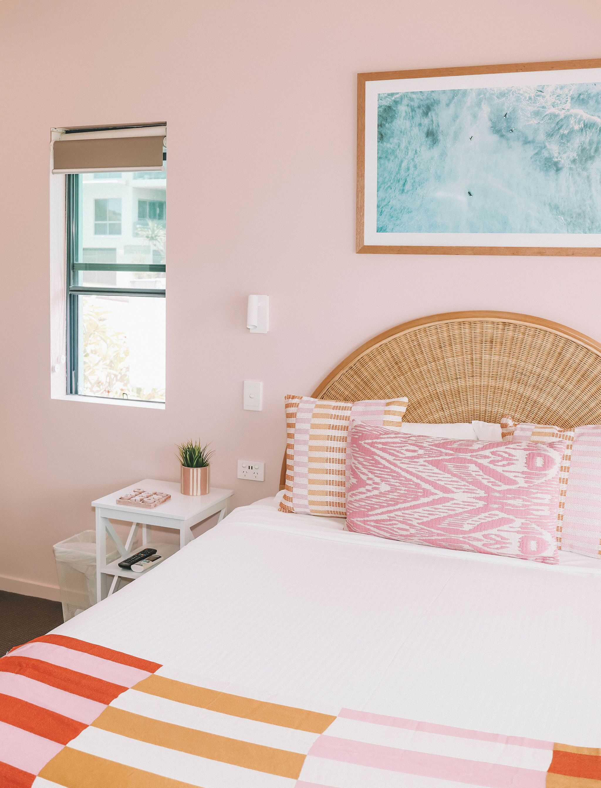 A bedroom with a bed, a bedside table with a plant, a window, and a painting on the wall.