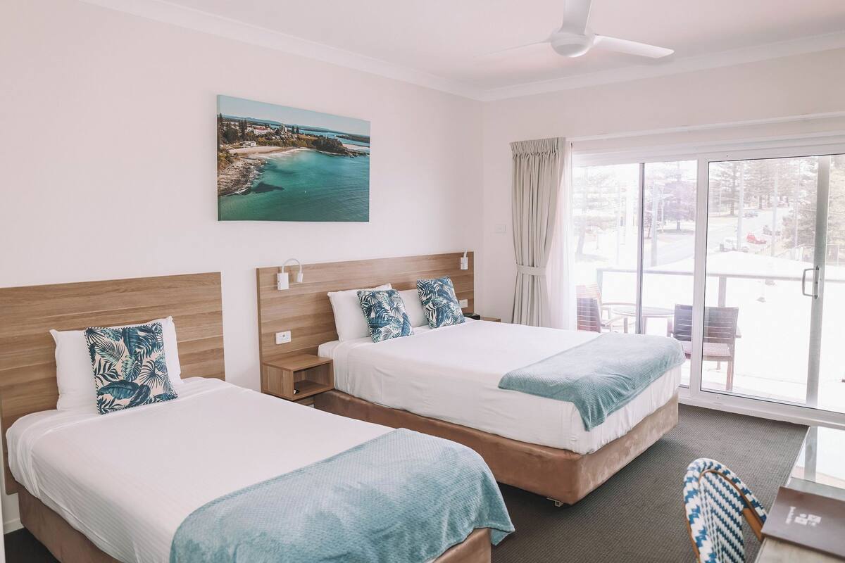 A hotel room with two beds, a painting of a coastal scene, a ceiling fan, and a view of the outdoors through a glass door.