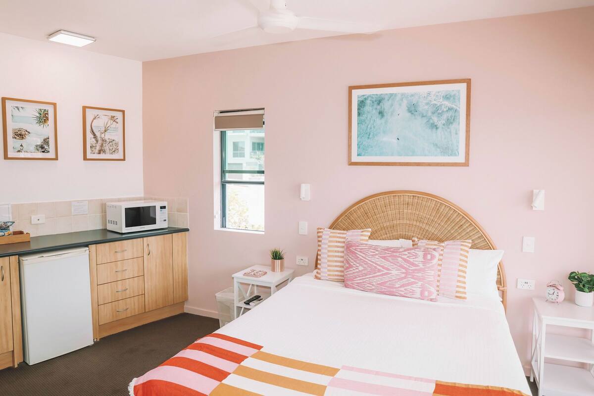 A bedroom with a bed, a bedside table, a nightstand, a microwave, a refrigerator, a window, and artwork on the walls.
