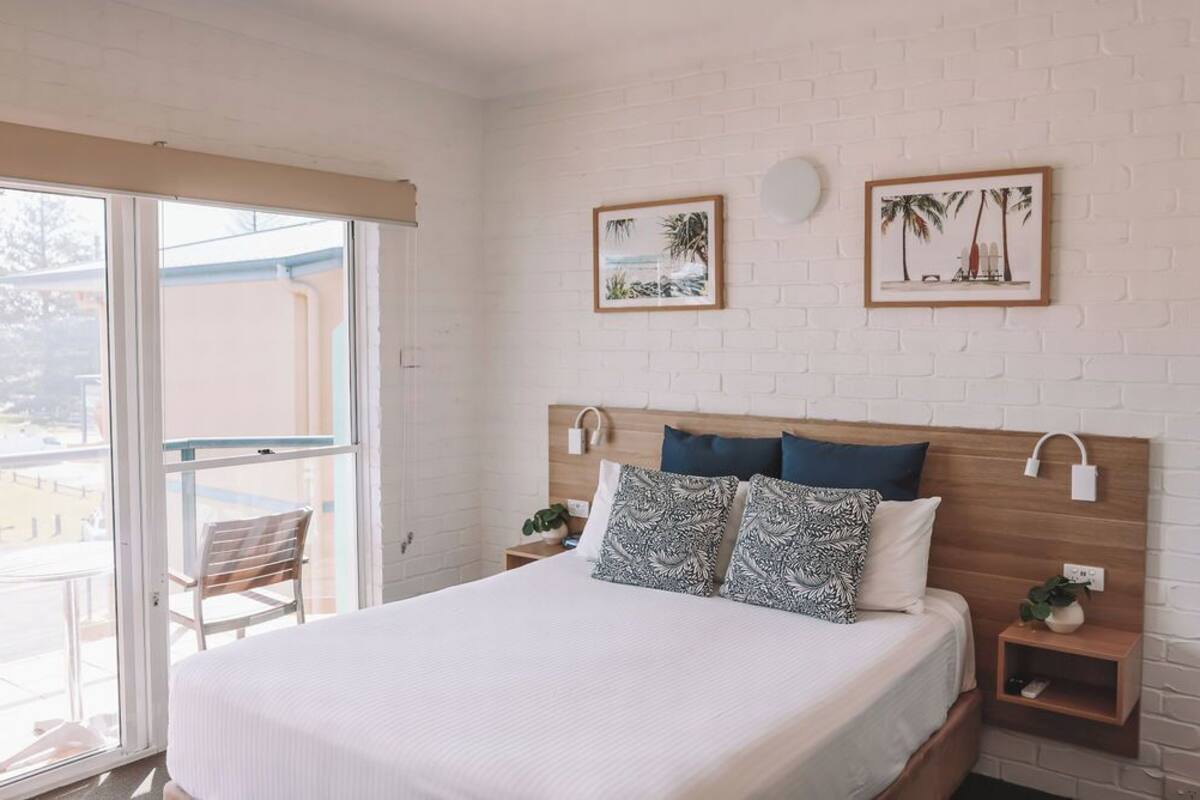 A hotel room with a bed, two nightstands, a wooden headboard, a balcony with a chair, and two framed pictures on the wall.