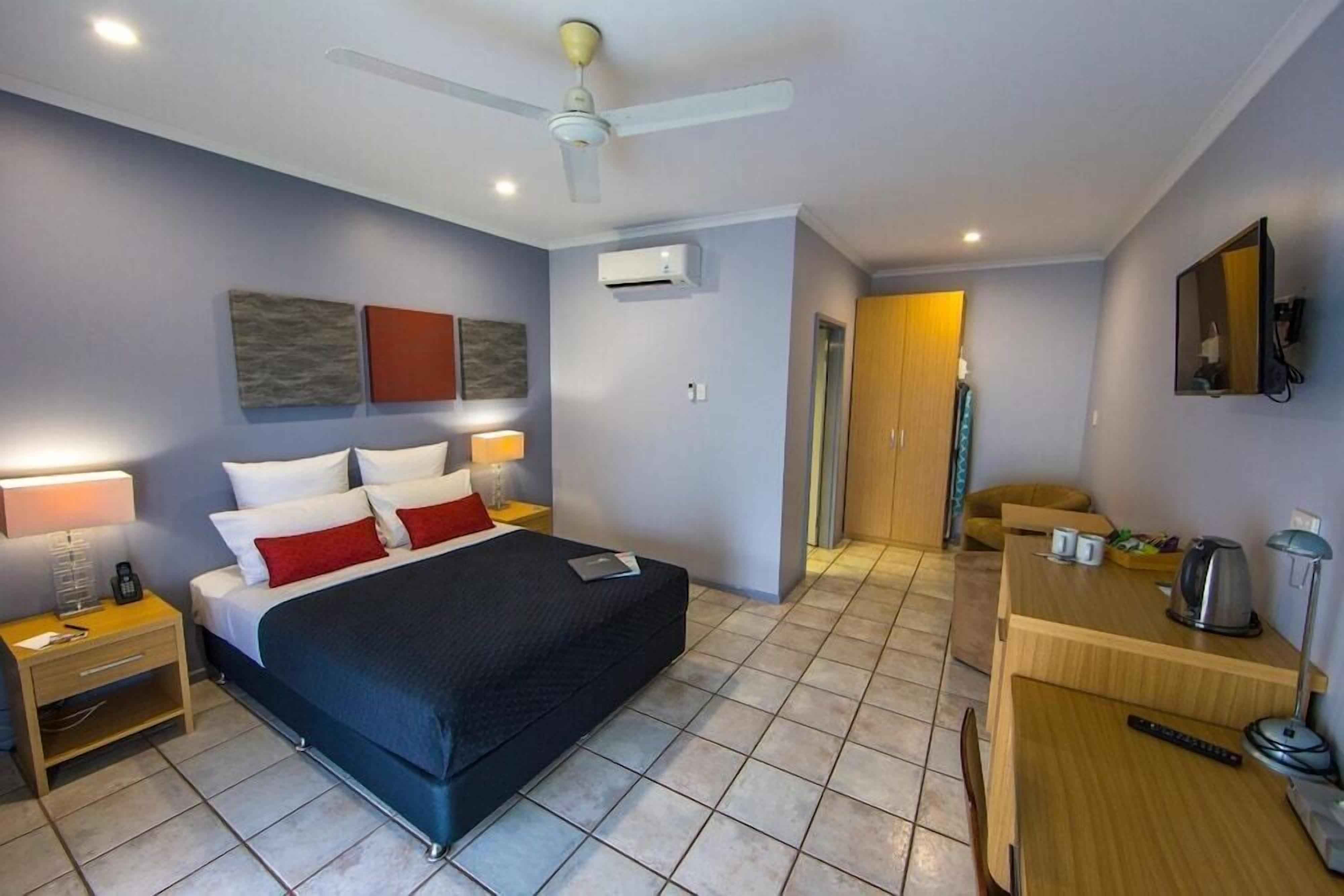 A hotel room with a bed, bedside table, lamp, air conditioning unit, and a desk with a chair.