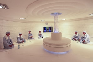 Ein Zimmer mit einer zentralen zylindrischen Struktur, einem Flachbildfernseher an der Wand und Personen, die im Kreis sitzen.