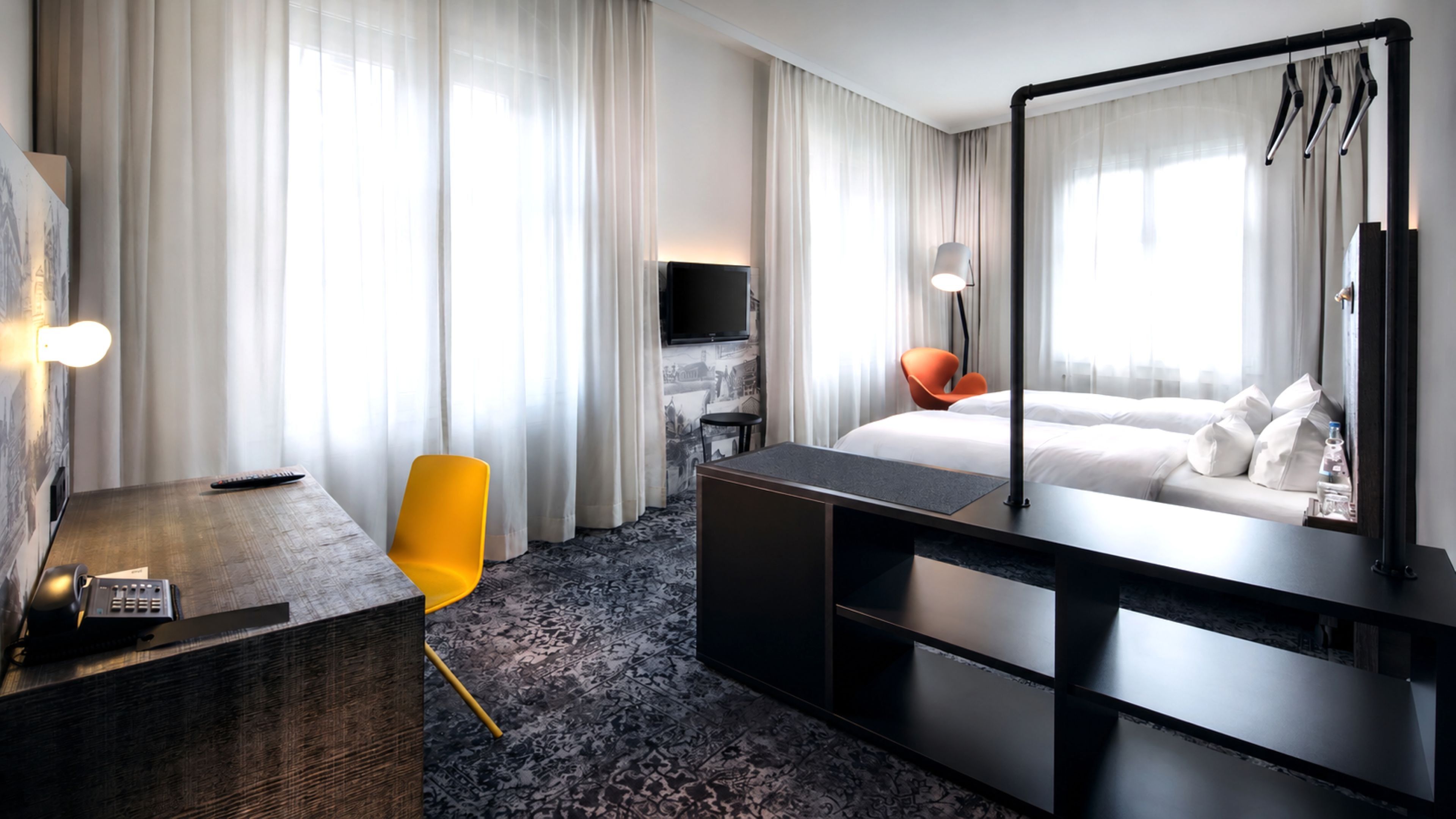 Habitación de hotel con cama, escritorio, silla, televisor y un amplio ventanal con cortinas.