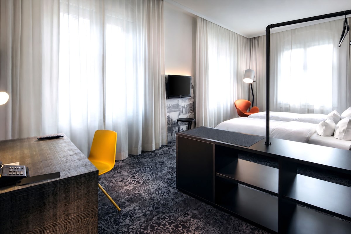 Habitación de hotel con cama, escritorio, silla, televisor y un amplio ventanal con cortinas.