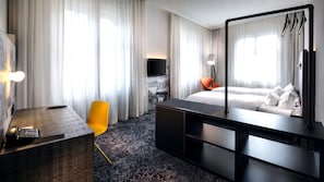Habitación de hotel con cama, escritorio, silla, televisor y un amplio ventanal con cortinas.