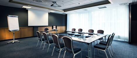 Un salón de conferencias con una pantalla de proyector, una pizarra y un amplio ventanal con persianas.