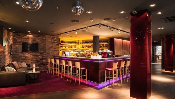 A bar area with a long counter, multiple stools, and a brick wall backdrop. - Pentahotel Braunschweig (Braunschweig)