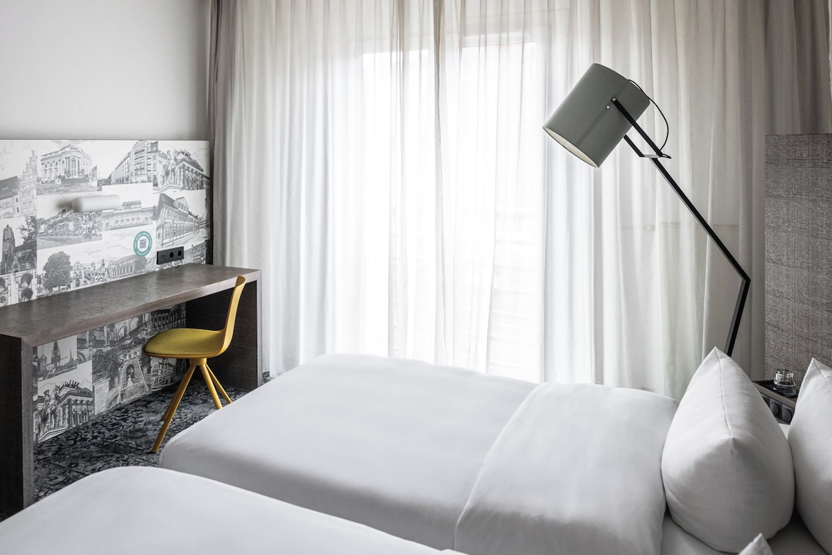 A hotel room with a bed, a desk, a chair, a TV, and a window with curtains.