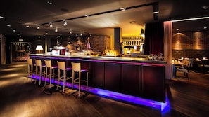 A bar area with a long counter, multiple stools, and a brick wall backdrop. - Pentahotel Braunschweig (Braunschweig)