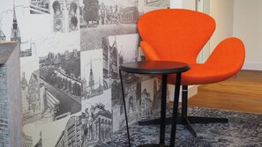 A room with a black round table and an orange chair, a patterned carpet, and a wall with black and white cityscape illustrations.
