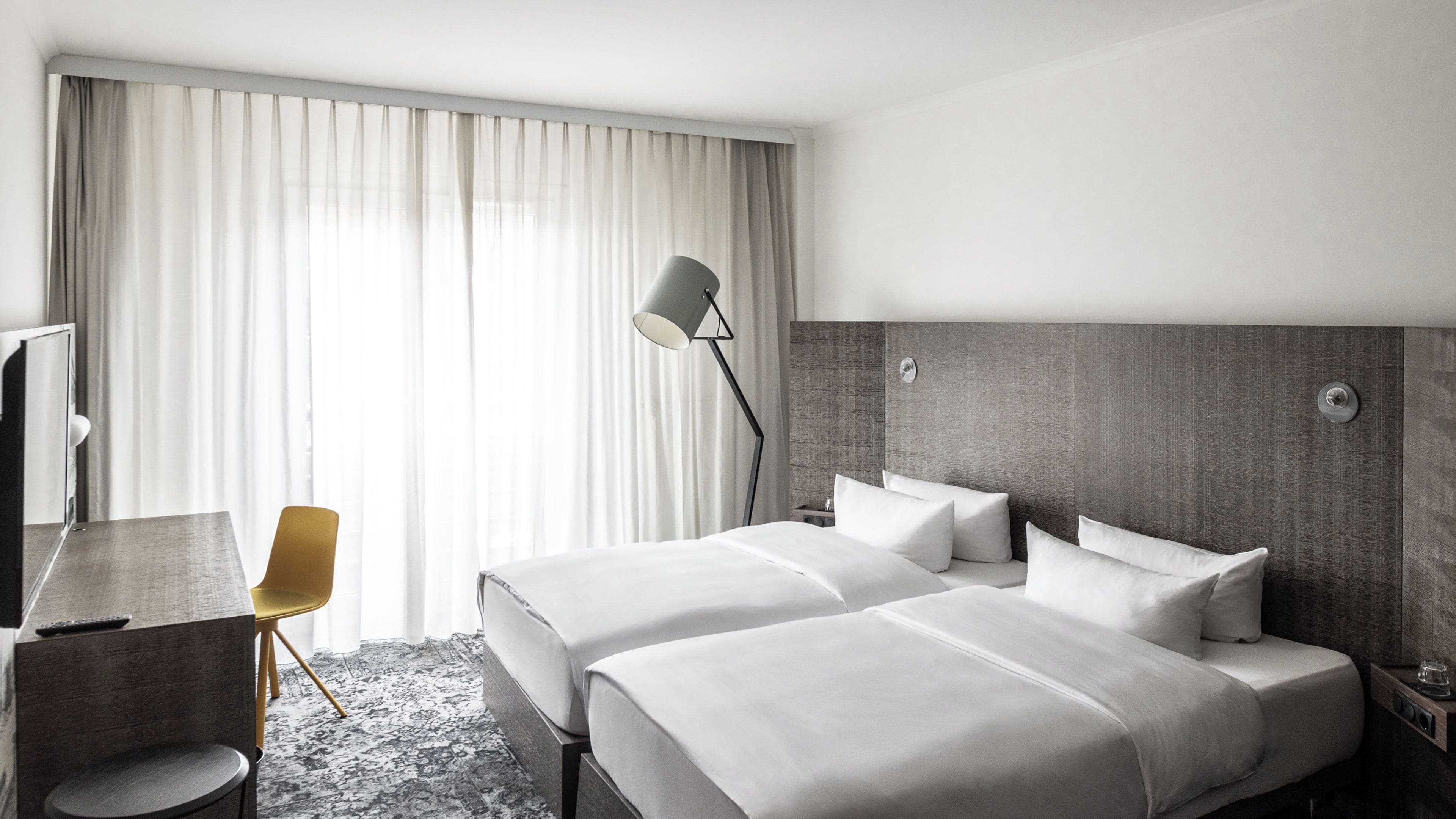 Habitación de hotel con dos camas, un escritorio, una silla, una lámpara y un ventanal con cortinas.