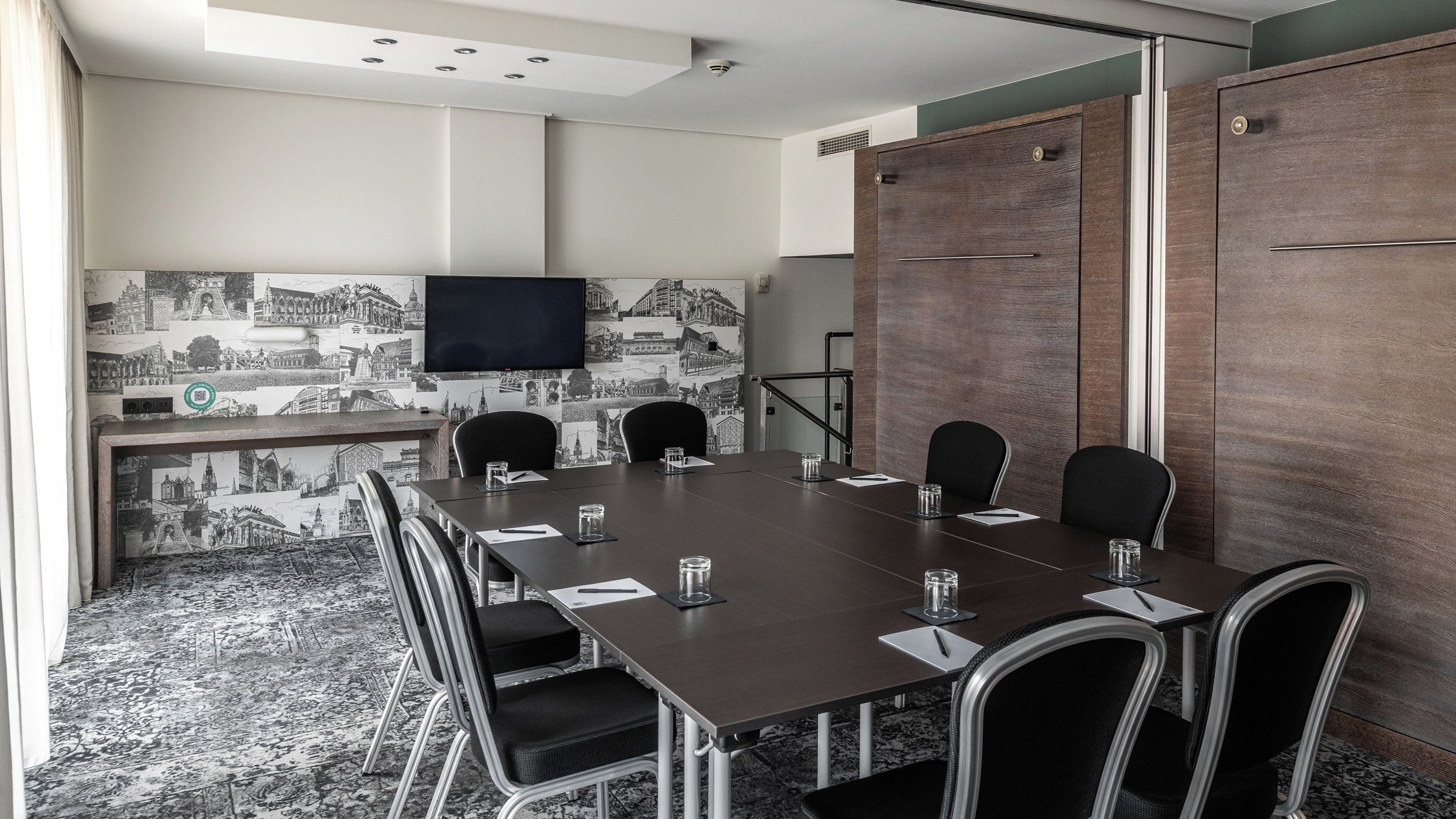 Un salón de conferencias con una larga mesa, sillas, un televisor de pantalla plana y un papel tapiz con vistas a la ciudad.