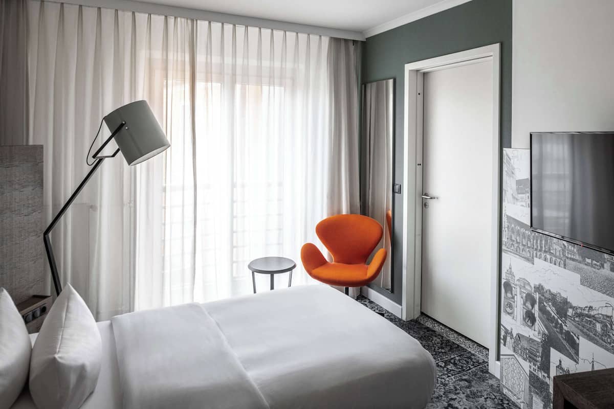Una habitación de hotel con cama, escritorio, silla, lámpara y un gran ventanal con cortinas.