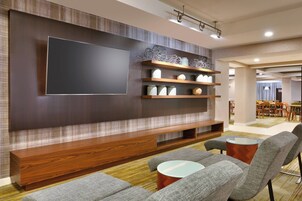 A modern living room with a large flat-screen TV, wooden shelves, and grey upholstered seating.