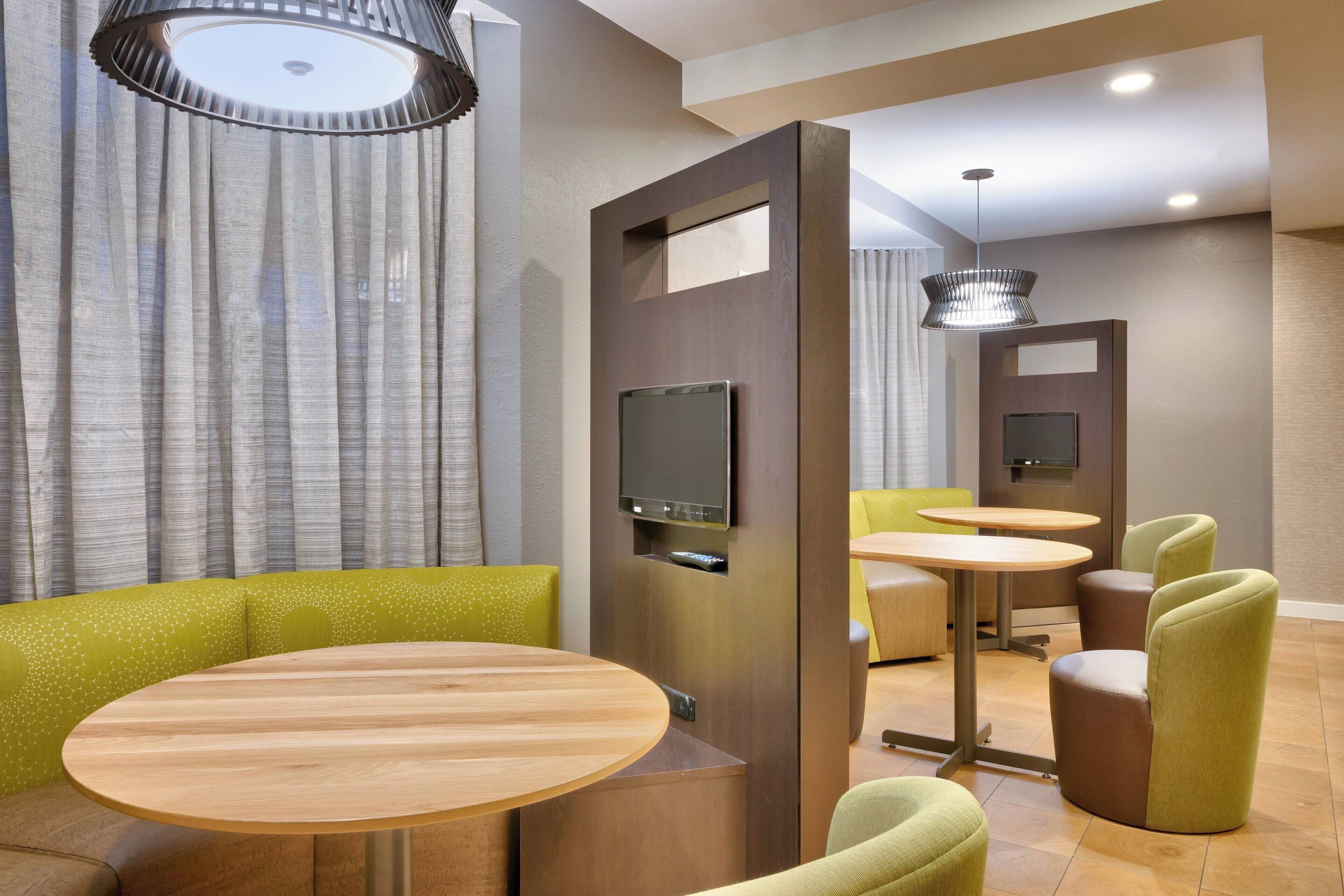 A modern hotel room with a wooden table, green upholstered seating, a flat-screen TV, and a ceiling light fixture.