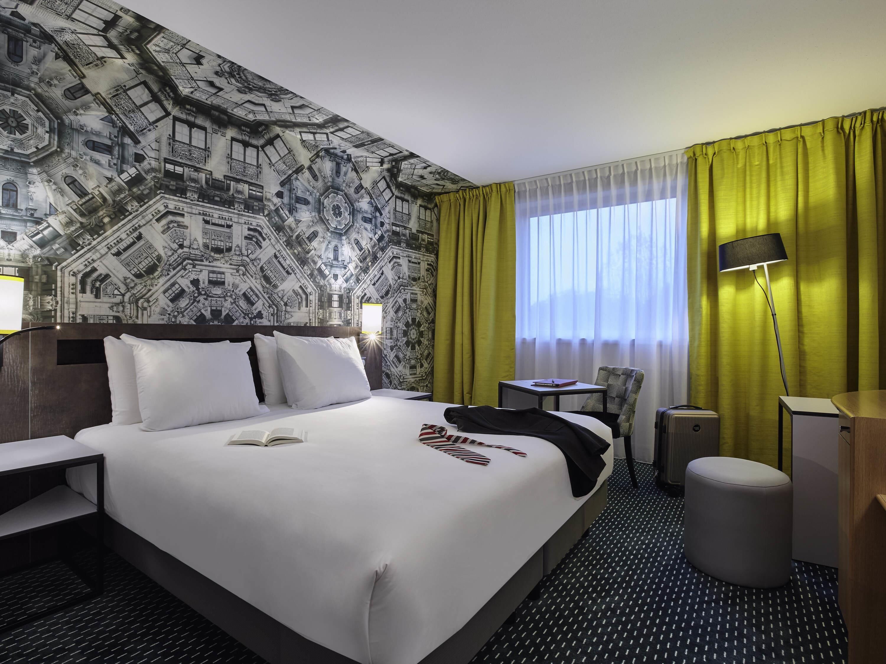A hotel room with a large bed, a desk, a chair, a lamp, and a view of the outside through a window with curtains.